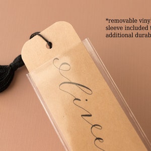 May include: A brown bookmark with the word "divine" written in black cursive script. The bookmark has a black tassel and is protected by a clear vinyl sleeve.