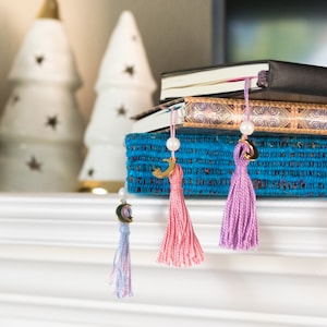 May include: Three books with gold and blue patterned covers are stacked on top of a blue box with a tassel bookmark hanging from each book. The tassels are pink, purple, and light blue.