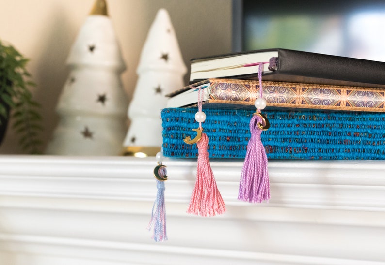 May include: Three books with gold and blue patterned covers are stacked on top of a blue box with a tassel bookmark hanging from each book. The tassels are pink, purple, and light blue.