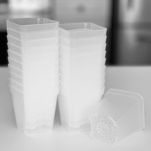 May include: A stack of clear, square-shaped plastic plant pots. The pots are white and have a ridged design. One pot is lying on its side, showing the drainage holes in the bottom. The background is blurred.