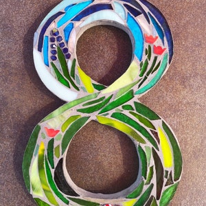 May include: A mosaic number eight made of glass tiles in shades of blue, green, and yellow. The number is surrounded by a border of green glass tiles. There are red and white mushroom accents at the bottom of the number.