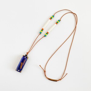 May include: A blue harmonica with the word "Swan" printed on it hangs from a brown cord necklace with white beads and colourful accents.