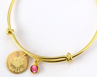 May include: Gold bangle bracelet with a charm that says "Charli" and has a paw print. The charm is attached to a small pink crystal.