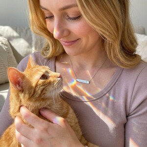 Woman wearing personalized paw print bar necklace while holding orange cat, custom pet memorial jewelry for cat lovers.