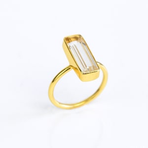 May include: A gold ring featuring a rectangular, clear gemstone. The gemstone is set in a gold bezel, and the ring has a simple, thin gold band. The ring is displayed on a white background, highlighting its elegant design.