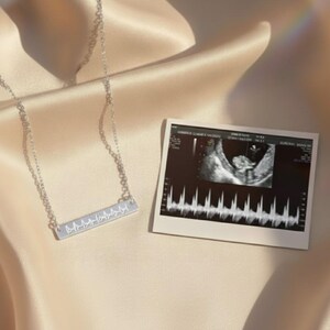 Sterling silver heartbeat bar necklace beside baby ultrasound print; custom fetal heartbeat soundwave engraving from sonogram, minimalist personalized pendant on adjustable chain; pregnancy announcement jewelry, push present, new mom gift.