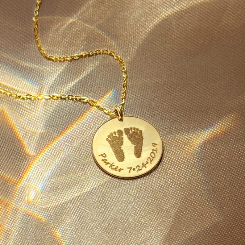 Close-up of personalized footprint necklace on champagne satin fabric, featuring engraved baby feet and name on a gold disc pendant. Available in sterling silver, gold filled, or rose gold filled with cable or satellite chain styles. A meaningful keepsake jewelry piece for new moms, baby shower gifts, push presents, or memorial remembrance necklaces honoring a child’s memory. Minimalist design ideal for everyday wear and sentimental gifting for baby loss gift, memorial present.