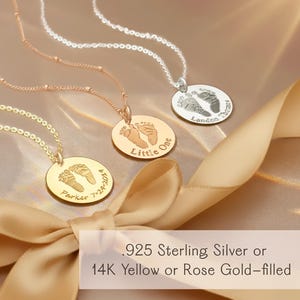 Three baby footprint necklaces in gold, rose gold, and silver hanging in warm sunlight near a window. Each round pendant features engraved footprints and custom text, showcasing personalized newborn keepsake jewelry in a soft, glowing setting.