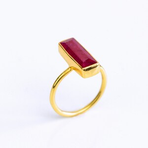 May include: A gold ring with a rectangular, deep red gemstone. The gemstone is set in a gold bezel, and the ring band is a simple, polished gold. The ring is set against a white background.
