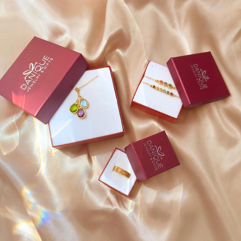 Elegant jewelry gift set displayed in branded Danique Jewelry gift boxes on a soft satin background, featuring a custom family birthstone necklace with peridot, aquamarine, and amethyst charms in gold bezels, a personalized gold engraved band ring, and a dainty gemstone bracelet with multiple stones. Perfect for Mother’s Day, anniversary, or bridesmaid gifts, this personalized jewelry collection highlights keepsake quality and thoughtful packaging ready for gifting.