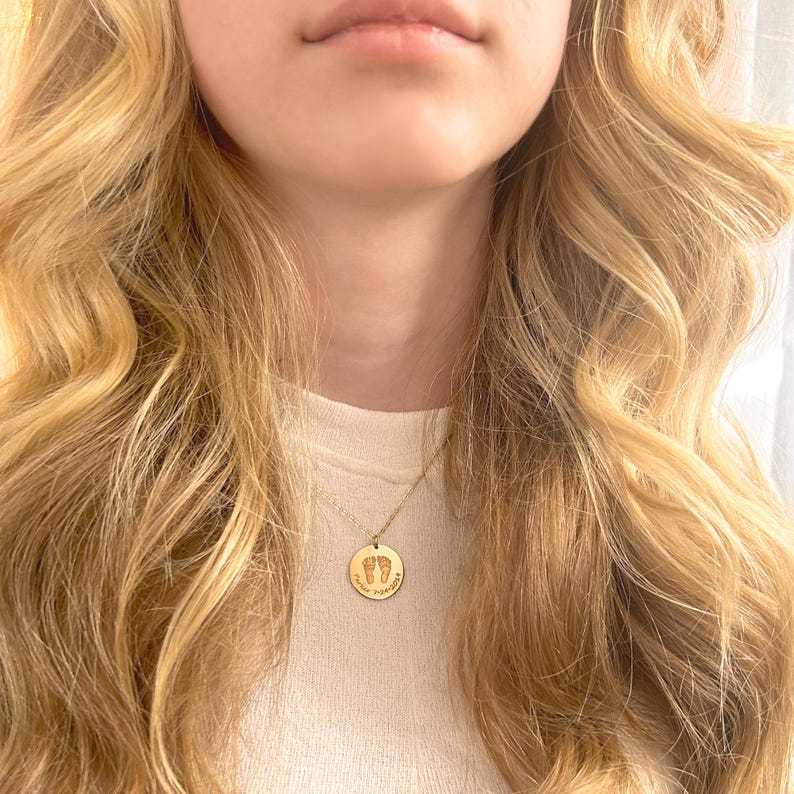 Model wearing a dainty personalized footprint necklace with engraved baby feet and custom name on a gold pendant. This minimalist everyday necklace is crafted in sterling silver, 14K gold filled, or rose gold filled, and can be customized with actual footprints or handprints sent by the customer. Perfect as a sentimental new mom necklace, baby shower keepsake, push present, grandmother jewelry gift, or remembrance necklace honoring a baby’s memory. A meaningful piece designed for daily wear love