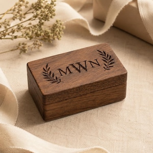 May include: A rectangular dark brown wooden box with a hinged lid. The top of the lid features an engraved design consisting of the letters MWN centered between two symmetrical laurel leaf branches, all etched into the wood grain surface.