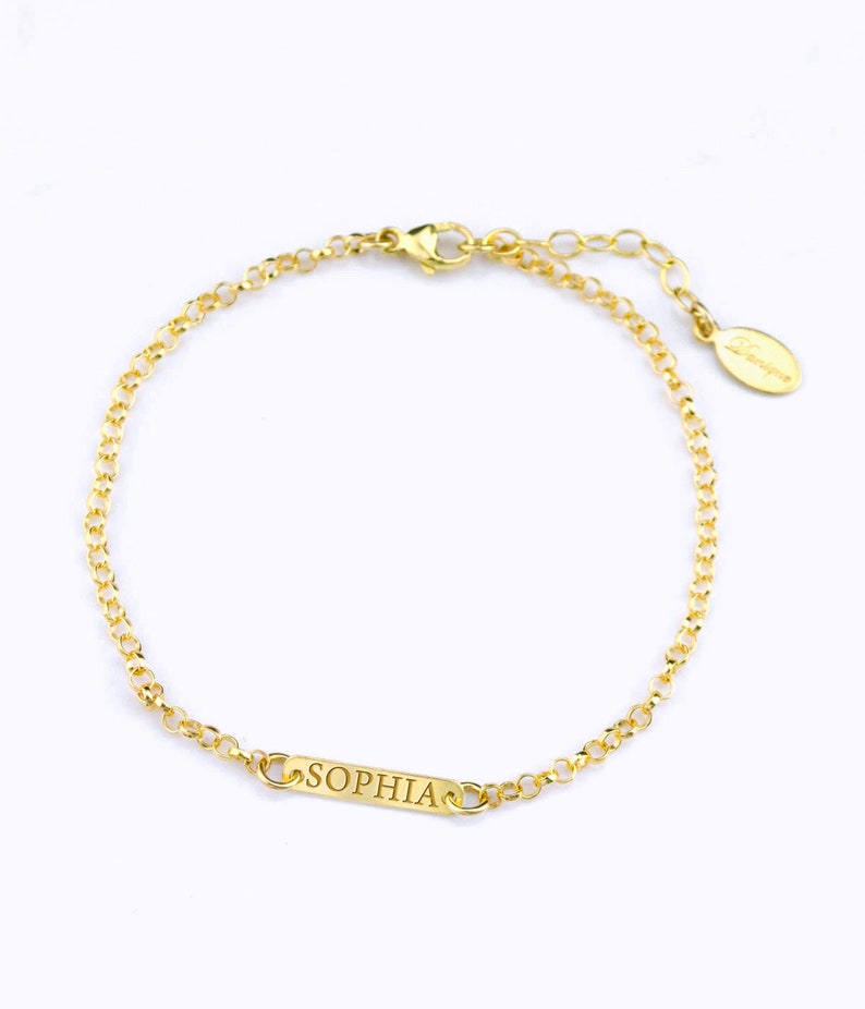 Puede incluir: Un brazalete de cadena dorada con una peque&ntilde;a etiqueta rectangular que dice "SOPHIA" grabada en ella.