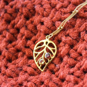May include: A gold leaf pendant necklace with two gemstones. The larger gemstone is red and the smaller gemstone is yellow. The necklace is on a red crocheted surface.