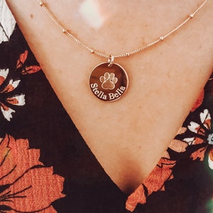 A rose gold paw print necklace with Stella Bella engraved in Classic font on a satellite chain. Pet necklace, custom pet jewelry, personalized pet jewelry, rose gold necklace with engraved pendant, custom engraved disc necklace