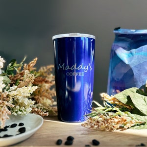 A blue coffee mug engraved with the phrase Maddys Coffee sits on a wooden table. A coffee bag is blurry in the background and coffee beans can be seen in the foreground. Gift for mom, mothers day gift for coffe lover mom, birthday gift for mom