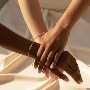 dainty personalized tiny bar bracelets in 18k gold vermeil worn by two friends with different skin tones, hands gently stacked on soft champagne satin fabric, each slim nameplate bar engraved with custom text and attached to a delicate adjustable chain, minimalist gold bracelet style perfect for everyday wear, friendship bracelets, bridesmaid gifts or matching mother daughter jewelry, customizable monogram or name bracelet handcrafted in sterling silver or gold vermeil