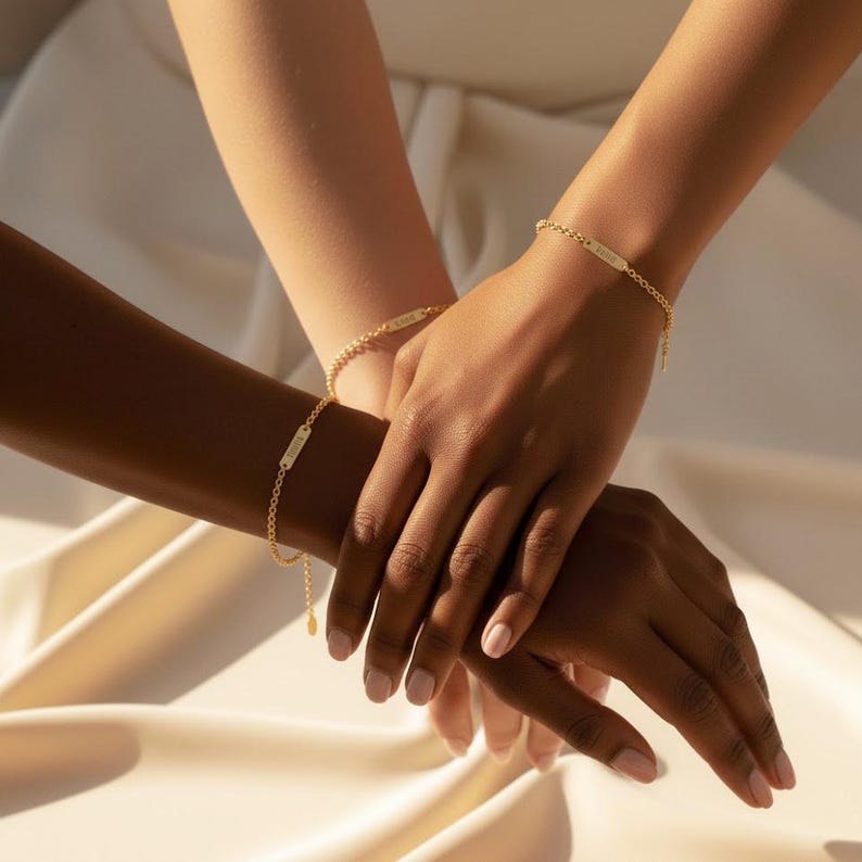 dainty personalized tiny bar bracelets in 18k gold vermeil worn by two friends with different skin tones, hands gently stacked on soft champagne satin fabric, each slim nameplate bar engraved with custom text and attached to a delicate adjustable chain, minimalist gold bracelet style perfect for everyday wear, friendship bracelets, bridesmaid gifts or matching mother daughter jewelry, customizable monogram or name bracelet handcrafted in sterling silver or gold vermeil