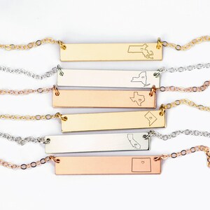 May include: Five gold, silver, and rose gold bar necklaces with state outlines and a heart in the center. The states are Maine, New York, Texas, North Dakota, California, and Colorado.