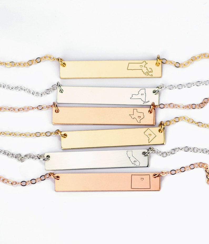 May include: Five gold, silver, and rose gold bar necklaces with state outlines and a heart in the center. The states are Maine, New York, Texas, North Dakota, California, and Colorado.