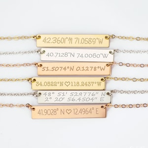 May include: Five gold, silver, and rose gold necklaces with bar pendants engraved with latitude and longitude coordinates. The pendants are engraved with different coordinates, including 42.3601&deg;N 71.0589&deg;W, 40.7128&deg;N 74.0060&deg;W, 51.5074&deg;N 0.1278&deg;W, 34.0522&deg;N 118.2437&deg;W, 48&deg; 51' 52.9776" N 2&deg; 20' 56.4504" E, and 41.9028&deg; N 12.4964&deg; E.