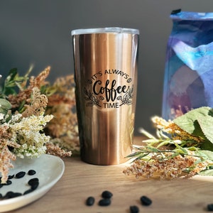 A copper toned stainless steel coffee tumbler engraved with a Its Always Coffee Time floral design engraved on the front sits on a wooden table with a coffee bag in the background and scattered coffee beans in the foreground. Gift for coffee lover