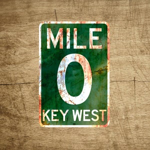May include: A green and white metal sign with the text "MILE 0 KEY WEST" on a wooden background.
