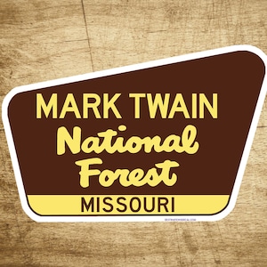 May include: A brown and white sign with the text "Mark Twain National Forest Missouri" in yellow letters. The sign has a white border and a rounded edge.