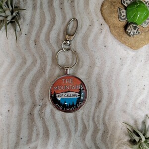 Może przedstawiać: Srebrny brelok do kluczy z okrągłym, pomarańczowym, niebieskim i białym breloczkiem z napisem "The Mountains Are Calling And I Must Go."