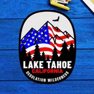May include: A vintage-style graphic design featuring a mountain range with an American flag draped over the peaks. The text "LAKE TAHOE CALIFORNIA DESOLATION WILDERNESS" is printed in a black circle around the mountains.