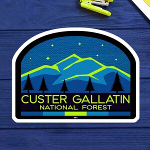 May include: A sticker featuring a mountain landscape with pine trees under a starry night sky. The text "CUSTER GALLATIN NATIONAL FOREST" is displayed in a bold, sans-serif font, with a black border.