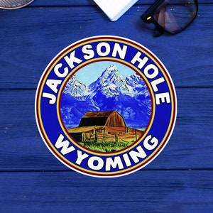 May include: A blue and gold circular sticker with the text "JACKSON HOLE WYOMING" around the edge. The center of the sticker features a mountain range with a barn in the foreground.