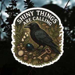 May include: A round sticker featuring a black crow with the text "SHINY THINGS ARE CALLING". The crow is surrounded by acorns, a key, and other natural elements. The sticker has a white border and a dark background.