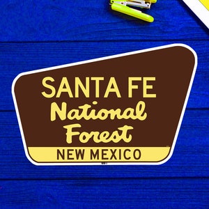 May include: A brown and yellow sign with the text "Santa Fe National Forest" and "New Mexico" on a blue wooden background.