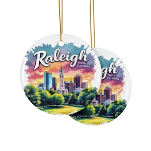 May include: A round ceramic ornament with a watercolor-style image of the Raleigh skyline. The ornament features a colorful sunset sky, green trees, and the text "Raleigh, North Carolina".