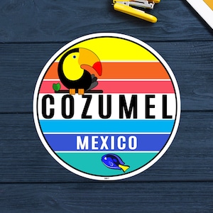 Op de afbeelding: Een ronde sticker met een kleurrijk ontwerp. Het toont een toekan boven het woord "COZUMEL" in het wit, met "MEXICO" eronder. De achtergrond heeft horizontale strepen in geel, oranje, rood en blauw. Onderaan een kleine vis.
