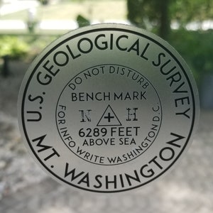 May include: A silver circular metal marker with black text that reads "U.S. Geological Survey" and "MT. WASHINGTON". The marker also includes the text "DO NOT DISTURB", "BENCH MARK", "FOR INFO WRITE WASHINGTON D.C.", and "6289 FEET ABOVE SEA".