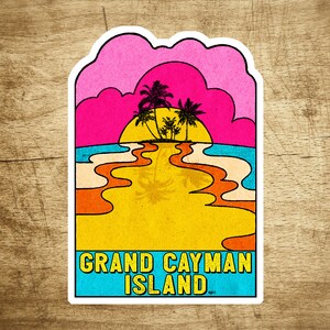 Puede incluir: Una pegatina de estilo retro con una escena de puesta de sol sobre una playa con palmeras. El texto "GRAND CAYMAN ISLAND" está impreso en azul y blanco en la pegatina.