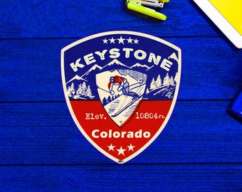 Könnte beinhalten: Ein vintage-artiger, rot-weiß-blauer Schildaufkleber mit den Worten "KEYSTONE Colorado" und "Elev. 10804 ft." Der Aufkleber zeigt einen Skifahrer in einem blau-roten Anzug, der einen Berg hinunterfährt.