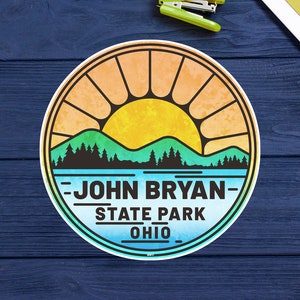 May include: A round sticker with a watercolor design of a sunset over a lake and mountains. The text "John Bryan State Park Ohio" is printed in black on the sticker.