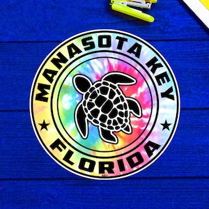 Puede incluir: Una pegatina de tie-dye colorida con una silueta de tortuga marina negra en el centro. La pegatina dice "Manasota Key Florida" en un círculo blanco alrededor de la tortuga.