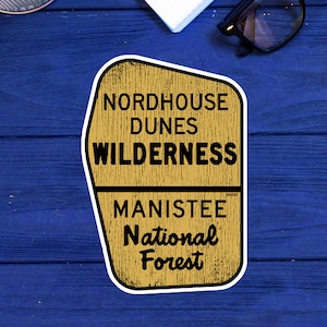May include: A brown and yellow sticker with the text "Nordhouse Dunes Wilderness Manistee National Forest" on a blue wooden background.