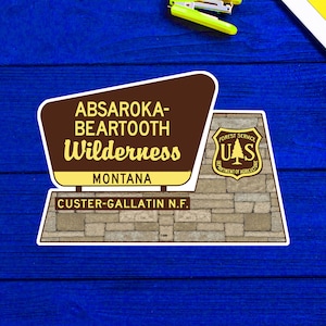 May include: A brown and yellow sign with the text "Absaroka-Beartooth Wilderness, Montana, Custer-Gallatin N.F." and a Forest Service emblem.