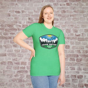 May include: A green t-shirt featuring a graphic of John Bryan State Park. The graphic includes a landscape scene with trees, a blue sky, and the park's name. The shirt is made of a soft material and is worn by a person.