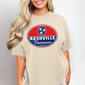 Op de afbeelding: Beige T-shirt met een rood en wit ovaal ontwerp met de tekst "NASHVILLE Tennessee" en drie sterren. Het shirt is een casual stijl.