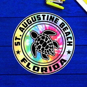 May include: A round sticker with a tie-dye background and a black silhouette of a sea turtle. The sticker says "St. Augustine Beach Florida" in a black circle around the turtle.