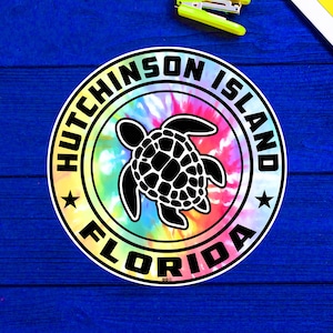 May include: A colorful tie-dye sticker with a black silhouette of a sea turtle. The sticker says "Hutchinson Island Florida" in a black circle.