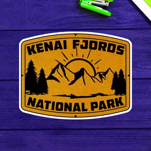 May include: A brown and yellow sticker with the text "Kenai Fjords National Park" in black lettering. The sticker features a stylized image of mountains, a sun, and trees.