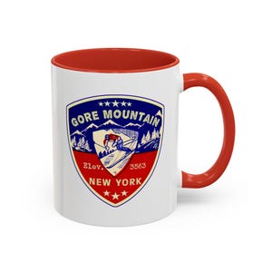 May include: White ceramic coffee mug with a red rim and handle. The mug features a vintage-style logo for Gore Mountain, a ski resort in New York. The logo is blue, red, and white and depicts a skier on a mountain.