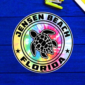 May include: A round sticker with a tie-dye background in shades of pink, yellow, blue, and green. The sticker features a black sea turtle illustration in the center, surrounded by a black ring with the words "JENSEN BEACH FLORIDA" and two black stars.